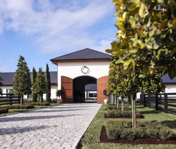 Entrance to the 3 Graces Dressage stables with a cobblestone pathway, manicured landscaping, and symmetrically planted trees under a blue sky