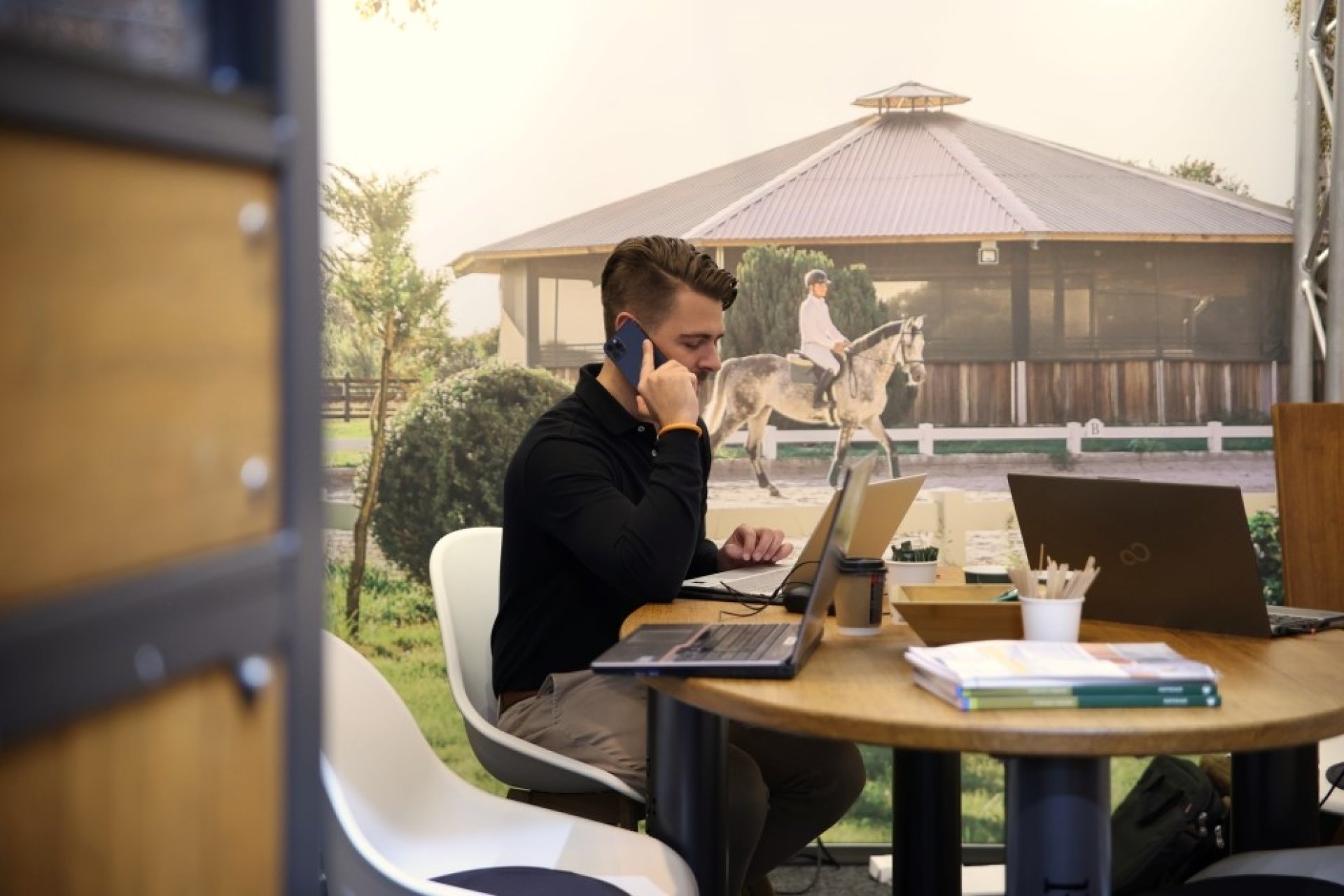 Fachinformatiker arbeitet im Büro an mehreren Laptops und telefoniert, im Hintergrund ein Reitstall mit Pferd und Reiter.